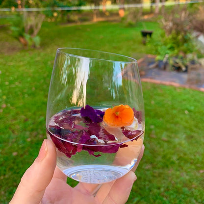 Clear glass with floral petals and ruby red Dulse floating in a gin cocktail held in front of a garden background