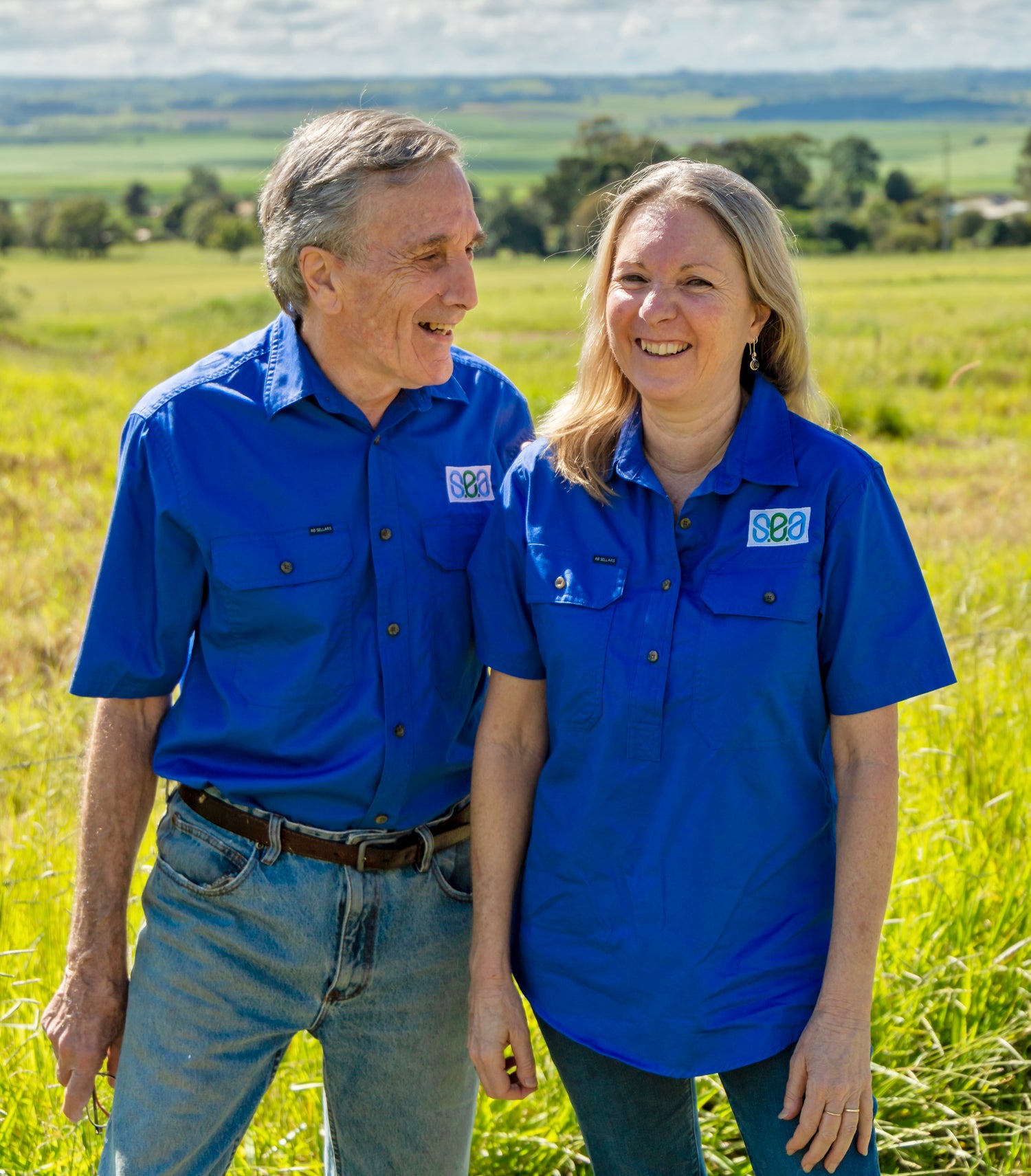 Grahame West and Liz Atkins standing in a grassy field with rolling farmland in the background.
